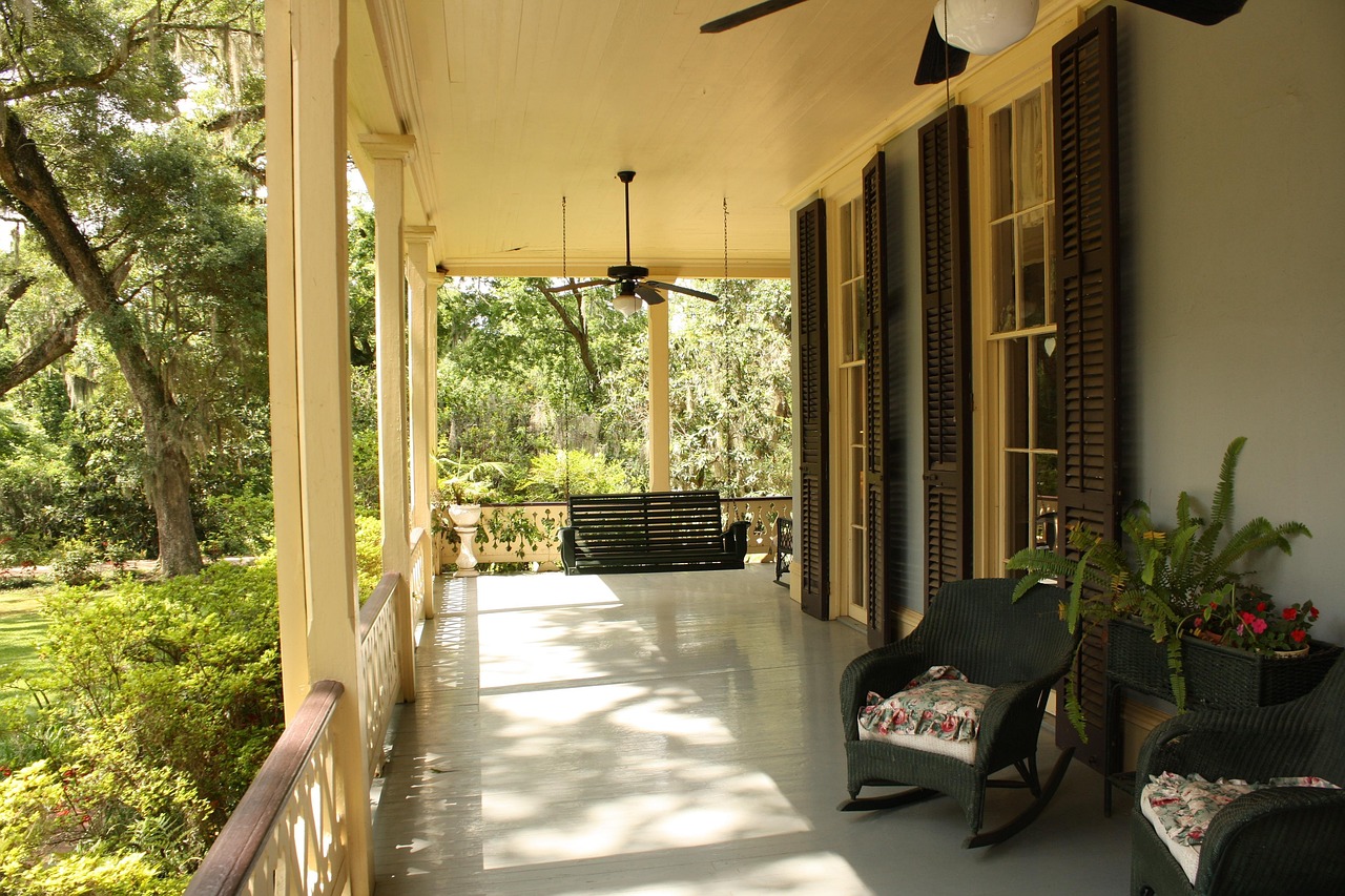 A bright, welcoming front porch with wicker rocking chairs, potted ferns, and a porch swing surrounded by lush greenery.