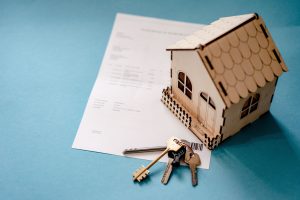 A small wooden house model and a set of keys resting on top of real estate paperwork against a soft blue background.