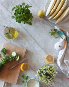 Fresh spring ingredients on a light wooden surface, including sliced cucumber, lemon, mint, yogurt, melon, and flowers arranged in a bright kitchen scene.